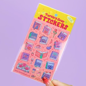 May include: A sheet of Turtle's Soup stickers featuring various book-themed illustrations on a pink background. The stickers include books labeled with romance tropes like Cowboy Romance, Sports Romance, and Enemies to Lovers, alongside small icons.