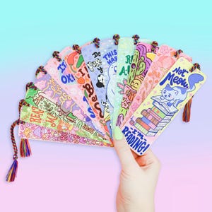 May include: A set of 10 colorful bookmarks with various illustrations and text. The bookmarks feature a variety of designs, including a cat reading a book, a stack of books, and a mushroom. The text on the bookmarks includes phrases like "I'm reading!" and "This has to be a dream".