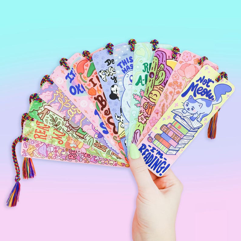 May include: A set of 10 colorful bookmarks with various illustrations and text. The bookmarks feature a variety of designs, including a cat reading a book, a stack of books, and a mushroom. The text on the bookmarks includes phrases like "I'm reading!" and "This has to be a dream".