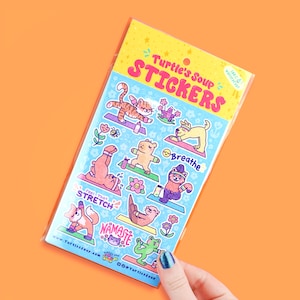 May include: A sheet of colorful stickers featuring various animals doing yoga poses. The stickers are on a blue background with white outlines. The text on the stickers includes "Turtle's Soup Stickers", "Breathe", "Feel That Stretch", and "Namaste".