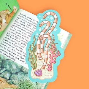 May include: A die-cut sticker featuring a light blue background with three tan and white striped garden eels emerging from sandy ground, surrounded by green seaweed, pink coral, and a small purple seashell, placed on an open book page.