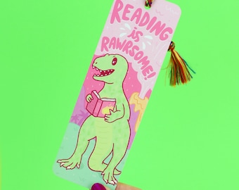 Puede incluir: Marcapáginas rosa y verde con un dinosaurio de dibujos animados sosteniendo un libro. El marcapáginas tiene el texto "Reading is Rawrsome!"