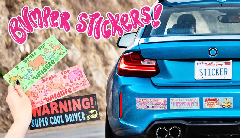 May include: A blue car with a variety of bumper stickers. The stickers feature illustrations of animals, a warning about a super cool driver, and a message about positivity. The license plate reads "Tintles Soup" and "STICKER".