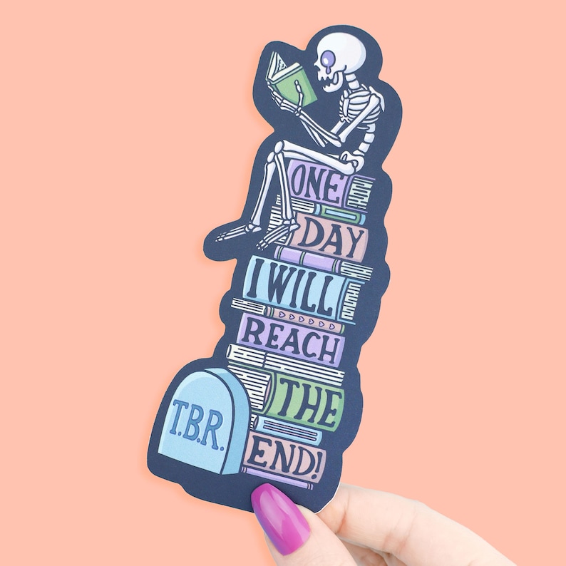 May include: A sticker showing a skeleton reading a book on a pile of books. The sticker has the text "ONE DAY I WILL REACH THE END!" and "T.B.R." on a tombstone. The sticker is held by a hand with a pink nail.