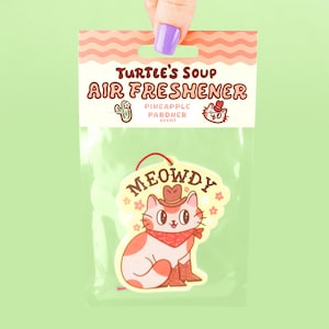 May include: A yellow and white car air freshener with a cartoon cat wearing a cowboy hat and bandana. The text "MEOWDY" is written in a yellow banner above the cat. The packaging says "Turtle's Soup Air Freshener Pineapple Pardner scent".