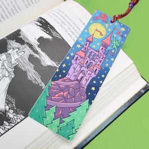 May include: A rectangular bookmark featuring a colorful illustration of a purple castle perched on a rocky cliff, surrounded by green pine trees, a yellow moon, and a small green dragon against a dark blue starry night sky, with a braided cord.