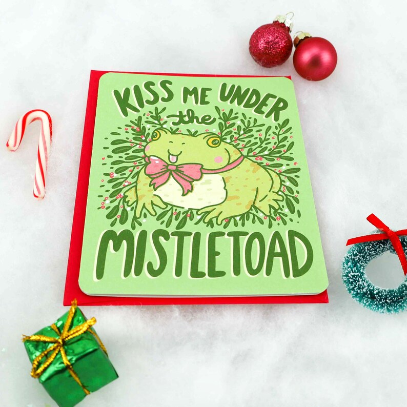 May include: A green Christmas card with a red border. The card features a cartoon frog with a pink bow tie and the text "Kiss me under the mistletoe".