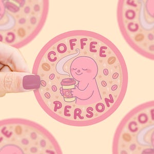 Puede incluir: Una pegatina redonda con borde rosa y las palabras "COFFEE PERSON" en rosa. Presenta una figura de dibujos animados rosa sosteniendo una taza de caf&eacute;, rodeada de granos de caf&eacute; y estrellas. El fondo es de color melocot&oacute;n claro.