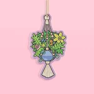 May include: A whimsical, illustrated air freshener in the shape of a hanging plant. The design features a blue pot filled with flowers, greenery, and a small snail. The macrame hanger is detailed with beads and a tassel, all on a lavender background.