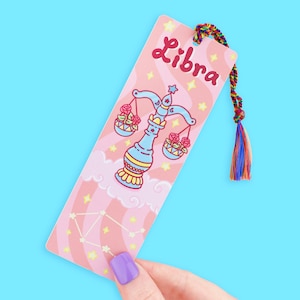 May include: A Libra-themed bookmark with a pink background, featuring a scale illustration and the word "Libra" in red. The bookmark has a colorful tassel and a constellation design.