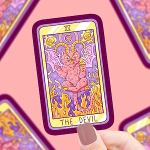 May include: A tarot card sticker featuring a pink cat-like devil with horns, wings, and a torch, surrounded by flames and skulls. The card is labeled "THE DEVIL" in white text against a purple border and a yellow background.