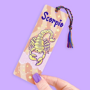 May include: A bookmark featuring a yellow scorpion illustration with the word "Scorpio" in purple. The bookmark has a colorful tassel and a background of stars and clouds. The bookmark is held by a hand with purple nail polish.