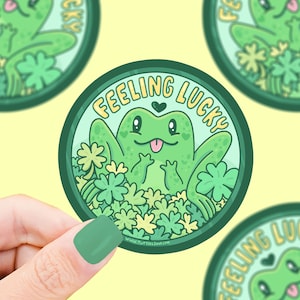 May include: A green frog with a big smile and tongue sticking out, surrounded by four-leaf clovers. The frog is sitting on a light blue background with the text "Feeling Lucky" in yellow letters.