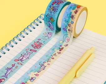 May include: Two rolls of washi tape with a beach theme. The tape features a variety of sea creatures, including sharks, narwhals, and jellyfish. The tape is on a white notebook with yellow lines.