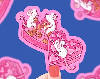 May include: Two white mice are sharing a pink heart-shaped box filled with chocolates. The box is decorated with small pink hearts. The mice are cartoon-style and have big eyes and smiles.