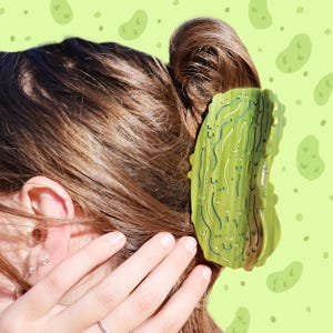 May include: A green hair clip shaped like a pickle is secured in brown hair. The clip has a textured surface with dark green and white details. The background is light green with pickle-shaped designs.