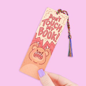 May include: A rectangular bookmark with the text "DON'T TOUCH MY BOOK!" and a cartoon bear illustration. The bookmark has a colorful braided tassel and is held by a hand with a purple painted nail.