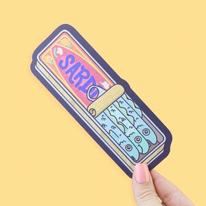 May include: A sticker featuring a cartoon illustration of a sardine can. The can is rectangular with a pink and yellow label that reads "SARIO" in blue. Inside are blue fish with cartoon eyes. The sticker has a dark blue border.