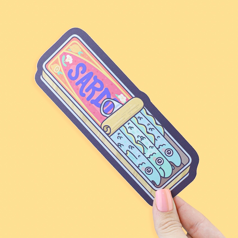 May include: A sticker featuring a cartoon illustration of a sardine can. The can is rectangular with a pink and yellow label that reads "SARIO" in blue. Inside are blue fish with cartoon eyes. The sticker has a dark blue border.