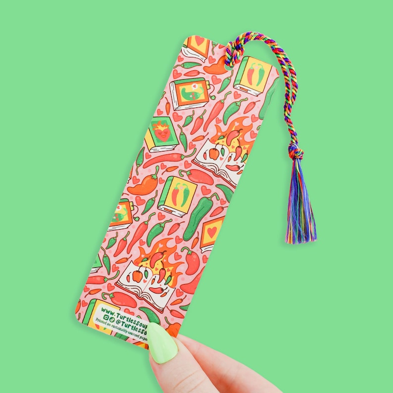 May include: A pink bookmark with a colorful pattern of chili peppers, hearts, and books. The bookmark has a tassel and the text "www.Turtlesout.co @Turtlesss" printed on the bottom.