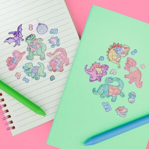 May include: Two notebooks, one lined and one solid mint green, with colorful dinosaur stickers. The stickers feature dinosaurs wearing sunglasses and musical notes. A green pen and a blue pen are also visible on a pink background.