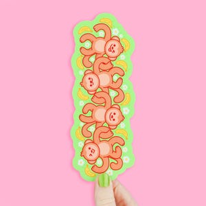 May include: A sticker featuring a stack of five cartoon monkeys in a row, each with a smiling face and orange fur. The monkeys are on a green background with bananas and white flowers, against a pink backdrop.