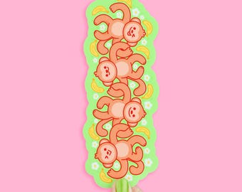 May include: A sticker featuring a stack of five cartoon monkeys in a row, each with a smiling face and orange fur. The monkeys are on a green background with bananas and white flowers, against a pink backdrop.