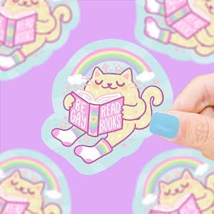 May include: A sticker featuring a cartoon cat reading a book with the text "BE GAY READ BOOKS". The cat is light yellow with a rainbow and clouds in the background. The sticker has a light blue border and is held by a hand with a blue fingernail.