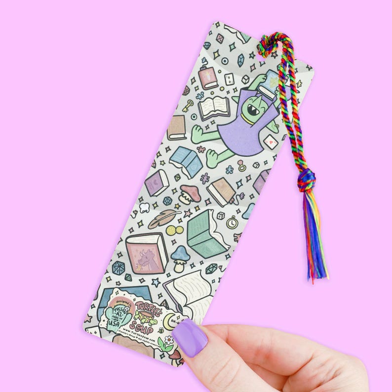 May include: A whimsical bookmark featuring a green alien in a purple robe, surrounded by books and magical elements. The bookmark has a rainbow tassel and is held by a hand with a purple nail polish.