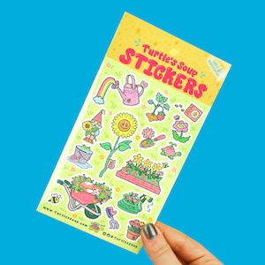 May include: A sheet of colorful stickers featuring a gardening theme. The stickers include a gnome, a sunflower, a watering can, a snail, a bee, a cat in a wheelbarrow, a garden hose, a trowel, a flower pot, and a seed packet. The stickers are on a light green background with a white border. The text "Turtle's Soup Stickers" is at the top of the sheet.