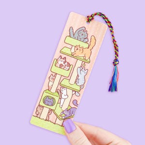 May include: A colorful bookmark featuring a cat tree design with various cartoon cats in different poses. The bookmark has a pink background with a rainbow tassel attached at the top. The bookmark is held by a hand with a purple painted nail.