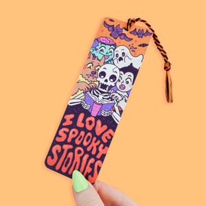 May include: A colorful bookmark featuring a skeleton reading a book with a group of spooky characters including a werewolf, a zombie, a vampire, and a ghost. The bookmark has the text "I love spooky stories" in a purple, orange, and red font.