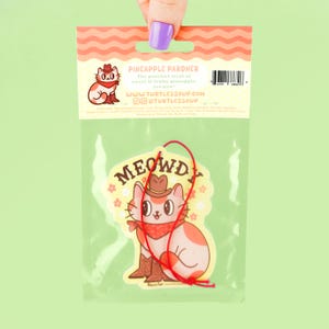 May include: A white car air freshener with a cartoon cat wearing a cowboy hat and bandana. The cat is sitting on a yellow background with pink flowers. The text "MEOWDY" is written in red letters above the cat. The back of the packaging says "Pineapple Pardner" and has a website address.