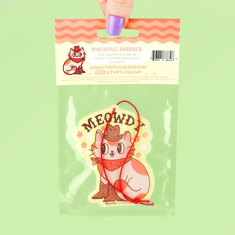 May include: A white car air freshener with a cartoon cat wearing a cowboy hat and bandana. The cat is sitting on a yellow background with pink flowers. The text "MEOWDY" is written in red letters above the cat. The back of the packaging says "Pineapple Pardner" and has a website address.