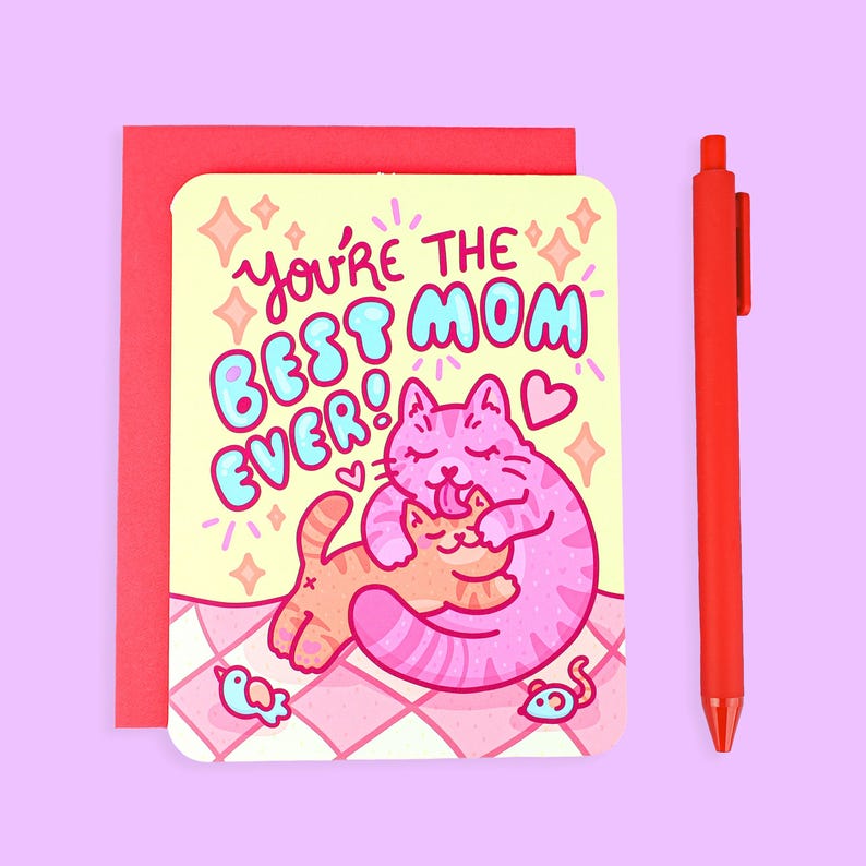 May include: A greeting card with the text "You're the Best Mom Ever!" features a pink cat hugging an orange cat. A red pen and envelope are next to the card. The card has a light yellow background with a pink and white checkered pattern.
