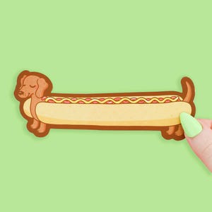 May include: A whimsical sticker featuring a dachshund dog cleverly designed as a hot dog. The dog's body forms the bun, with the head and tail at either end. The hot dog has mustard and ketchup toppings. The sticker is held by a hand with green nail polish.