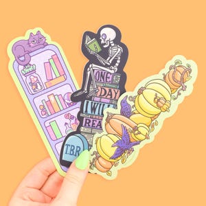May include: Three quirky stickers: a bookshelf with a purple cat, a skeleton reading on books, and pumpkins with a purple crow. The bookshelf sticker has the letters "T.B.R." on a tombstone. The skeleton sticker has the words "ONE DAY I WILL READ".