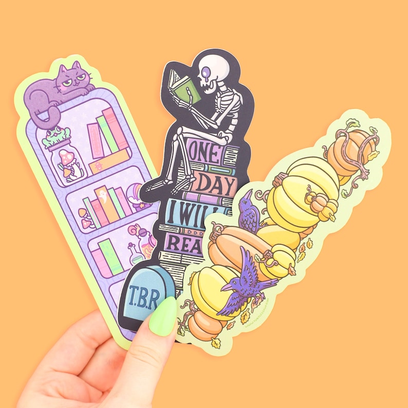 May include: Three quirky stickers: a bookshelf with a purple cat, a skeleton reading on books, and pumpkins with a purple crow. The bookshelf sticker has the letters "T.B.R." on a tombstone. The skeleton sticker has the words "ONE DAY I WILL READ".