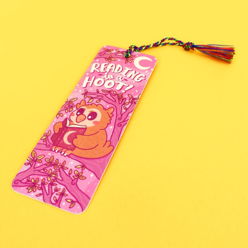 Pu&ograve; includere: Segnalibro rosa con un gufo dei cartoni animati che legge un libro con una luna crescente sulla copertina. Il segnalibro ha il testo "Reading is a Hoot!" e una luna crescente nella parte superiore.