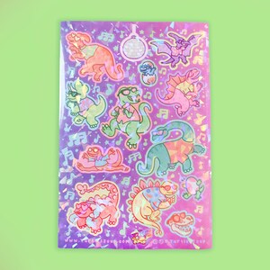 May include: A holographic sticker sheet featuring cartoon dinosaurs in sunglasses, musical notes, and a disco ball. The dinosaurs are in various poses and colors, including orange, green, and pink, on a purple background.
