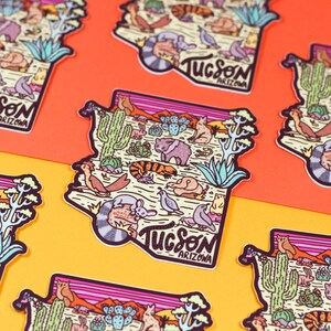 May include: A colorful sticker featuring an outline of the state of Arizona with the word "Tucson" and "Arizona" written inside. The sticker is filled with illustrations of desert animals and plants, including a cactus, a lizard, a bird, a scorpion, and a rabbit.