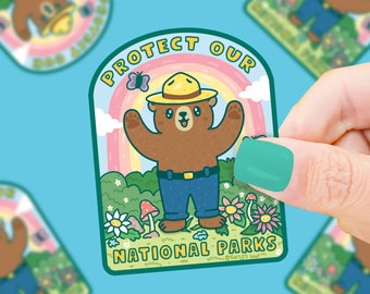 May include: A green, pink, and blue sticker with a cartoon bear wearing a ranger hat and blue jeans. The sticker says "Protect Our National Parks" with a rainbow and flowers in the background.