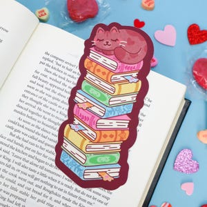 May include: A die-cut sticker featuring a stack of colorful books with a sleeping pink cat on top. The books are illustrated in shades of pink, orange, yellow, green, and blue with decorative patterns. The sticker is placed on an open book page.