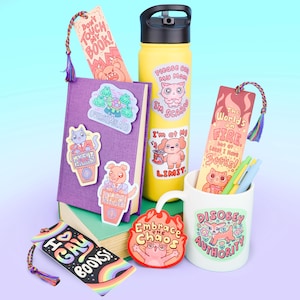 May include: A collection of stationery items, including bookmarks, stickers, and a mug. The bookmarks have colorful tassels and text like "I ❤️ GAY BOOKS!" and "The World's on FIRE but at Least I Have Books!". The stickers feature cartoon animals.