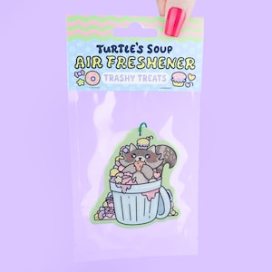 May include: Air freshener in a clear plastic package. The air freshener features a cartoon raccoon eating from a trash can overflowing with sweets. The packaging reads "Turtle's Soup Air Freshener Trashy Treats."