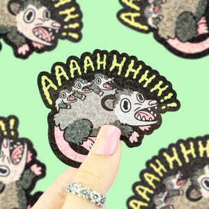 May include: A sticker featuring a cartoon opossum with the word "AAAAHHHHH!!" in yellow. The opossum is gray, white, and pink, with its mouth open in a scream. The sticker is held up by a hand with a pink nail.