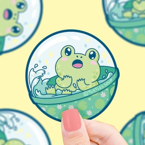 May include: A green frog with a surprised expression is inside a clear bubble with a blue wave and green lily pads. The bubble is surrounded by a green and white border with the text "TURTLE'S SOUP".