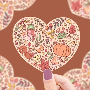 May include: A heart-shaped sticker with a whimsical autumn theme. It features a squirrel, a frog, a pumpkin, mushrooms, leaves, and other fall-themed elements in a colorful cartoon style.