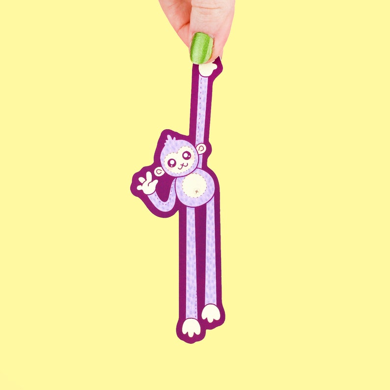 Puede incluir: Un mono morado con brazos y piernas largos est&aacute; agarrado a un dedo. El mono tiene una cara blanca con ojos negros y una nariz rosa. El mono est&aacute; sonriendo y tiene una barriga blanca.