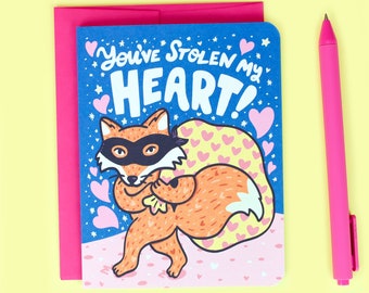 Op de afbeelding: Een blauwe kaart met een cartoonvos die een masker draagt en een tas met harten draagt. De kaart zegt "You've stolen my heart!" in witte letters.